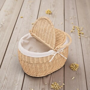 May include: Wicker basket with a hinged lid and a handle. The basket is oval-shaped and lined with white fabric. A white ribbon is tied on the front. The basket is light brown and sits on a wooden surface.