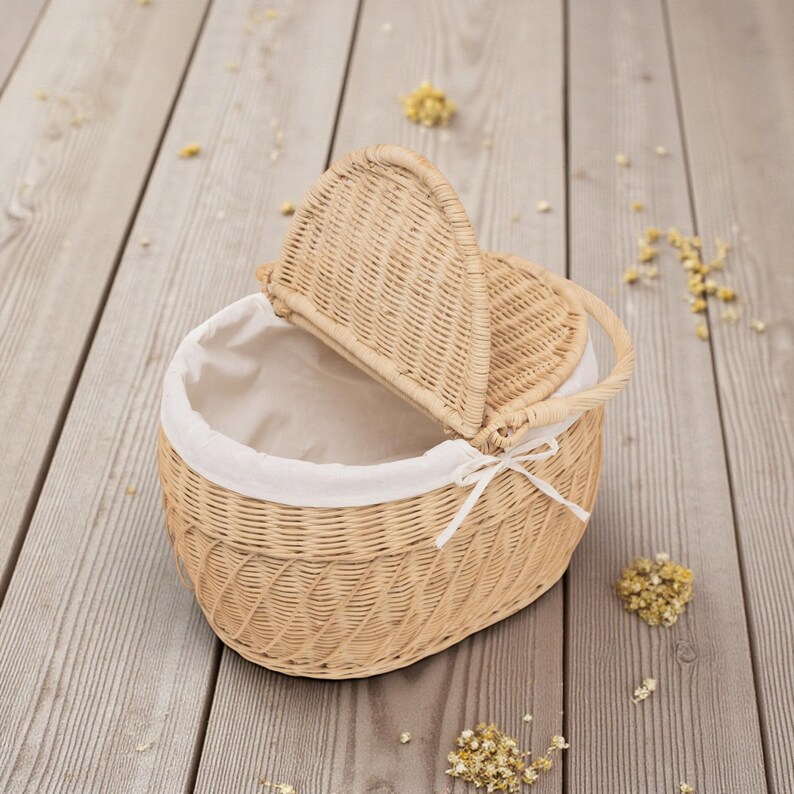 May include: Wicker basket with a hinged lid and a handle. The basket is oval-shaped and lined with white fabric. A white ribbon is tied on the front. The basket is light brown and sits on a wooden surface.