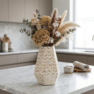 Pode incluir: Um vaso decorativo com um padrão geométrico contém um buquê de flores secas, algodão e capim-dos-pampas. O vaso é bege e branco, e as flores são em tons de marrom e creme. O arranjo está em uma bancada de mármore.