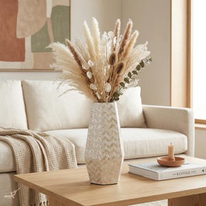Pode incluir: Um vaso geométrico alto com flores e ervas secas está sobre uma mesa de centro de madeira. O vaso tem um padrão de espinha de peixe em tons de creme e bege. O arranjo inclui capim-dos-pampas, vagens de sementes e eucalipto. Um sofá e uma obra de arte estão ao fundo.