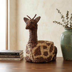 Puede incluir: Una cesta con forma de jirafa tejida en tonos marrones y beige. La jirafa tiene un cuello largo y un cuerpo redondo con un patrón de manchas. La cesta está hecha de materiales naturales y es adecuada para guardar o decorar.