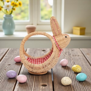 May include: A woven wicker Easter basket shaped like a bunny, with a red and white checkered lining. The basket sits on a wooden table, surrounded by speckled pastel-colored Easter eggs. A vase of yellow flowers is in the background.