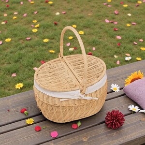 May include: A light brown wicker picnic basket with a handle and a hinged lid. The basket has a white fabric lining and is tied with a white ribbon. The basket is sitting on a wooden surface with flowers and petals scattered around.