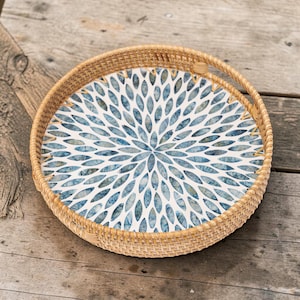Peut inclure: Plateau de service rond avec bord et poignée tressés. Le plateau présente un bord tressé marron clair et un intérieur blanc avec un motif floral bleu. Le plateau est posé sur une surface en bois.