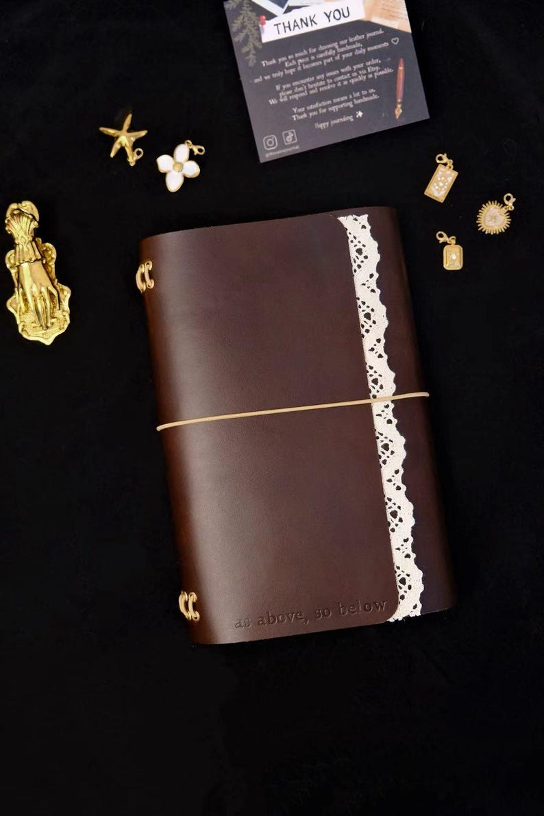 May include: A dark brown leather journal with a vertical white lace trim, secured by a thin elastic band. The cover features the embossed text 'as above, so below'. Surrounding the journal are gold-toned charms, including a hand, star, and flower.