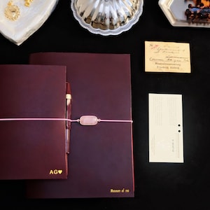 May include: Two dark burgundy leather journals with gold lettering. The smaller journal features the initials AG with a heart symbol, while the larger one is labelled Museum of me. Both are secured by a pink elastic band with a rectangular stone.