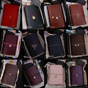 May include: A collection of twelve rectangular leather journals in various colours including red, black, brown, burgundy, mahogany, foresta, pastel pink, and purple. Each journal features a coloured elastic band with a unique metal charm attached.