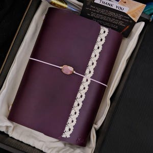 May include: A dark purple leather journal featuring a vertical strip of cream-coloured lace trim and a light pink elastic closure band secured with a central oval-shaped pink stone. The journal is presented in a black box with a thank you card.