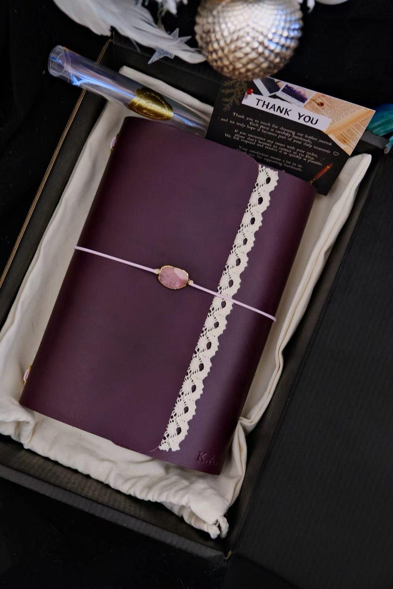 May include: A dark purple leather journal featuring a vertical strip of cream-coloured lace trim and a light pink elastic closure band secured with a central oval-shaped pink stone. The journal is presented in a black box with a thank you card.