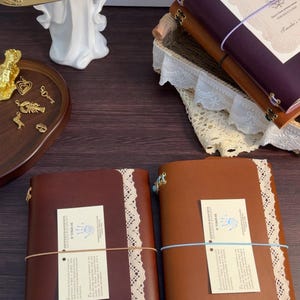 May include: Four leather journals in dark brown, tan, and deep purple, each featuring a white lace trim, elastic closure, and a certificate of authenticity. A gold tray holds small metal charms, including starfish and hearts, near a white angel figurine.