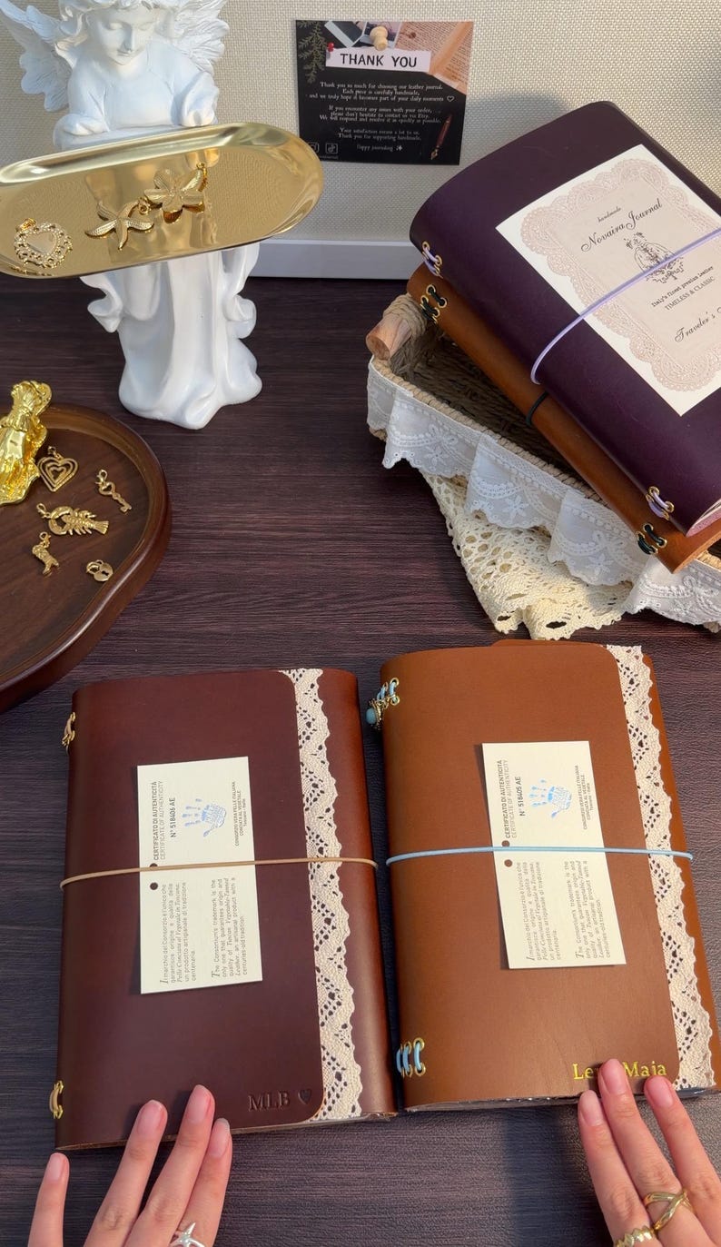 May include: Four leather journals in dark brown, tan, and deep purple, each featuring a white lace trim, elastic closure, and a certificate of authenticity. A gold tray holds small metal charms, including starfish and hearts, near a white angel figurine.