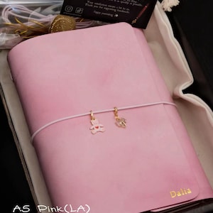 May include: A pink leather journal with a baby pink elastic cord featuring a gold bear charm and a crystal charm. The name Dalia is embossed in gold on the bottom right corner. A thank you card is included with the text Thank You and other details.