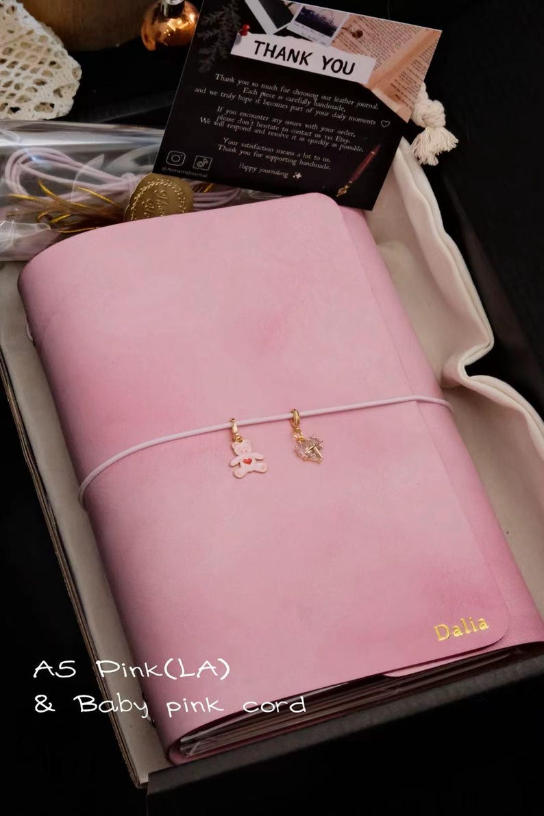 May include: A pink leather journal with a baby pink elastic cord featuring a gold bear charm and a crystal charm. The name Dalia is embossed in gold on the bottom right corner. A thank you card is included with the text Thank You and other details.