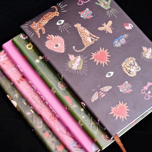 May include: A stack of notebooks with decorative covers. The top notebook is dark grey with leopard and heart illustrations. Other notebooks are pink, green, and floral patterned. A brown ribbon bookmark is visible.