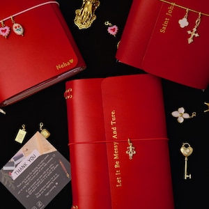 May include: Three red leather journals with elastic closures and gold foil text including Neha, Saint John 1:1-5, and Let It Be Messy And Ture. The journals are decorated with various gold-toned charms including hearts, a cross, a key, and a fleur-de-lis.