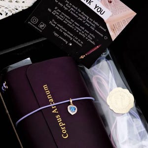 May include: A dark purple leather journal with the text Corpus Arcanum embossed in gold. A light purple elastic cord wraps around the journal, featuring a heart-shaped charm with a blue stone. A thank you card and extra cords are included.