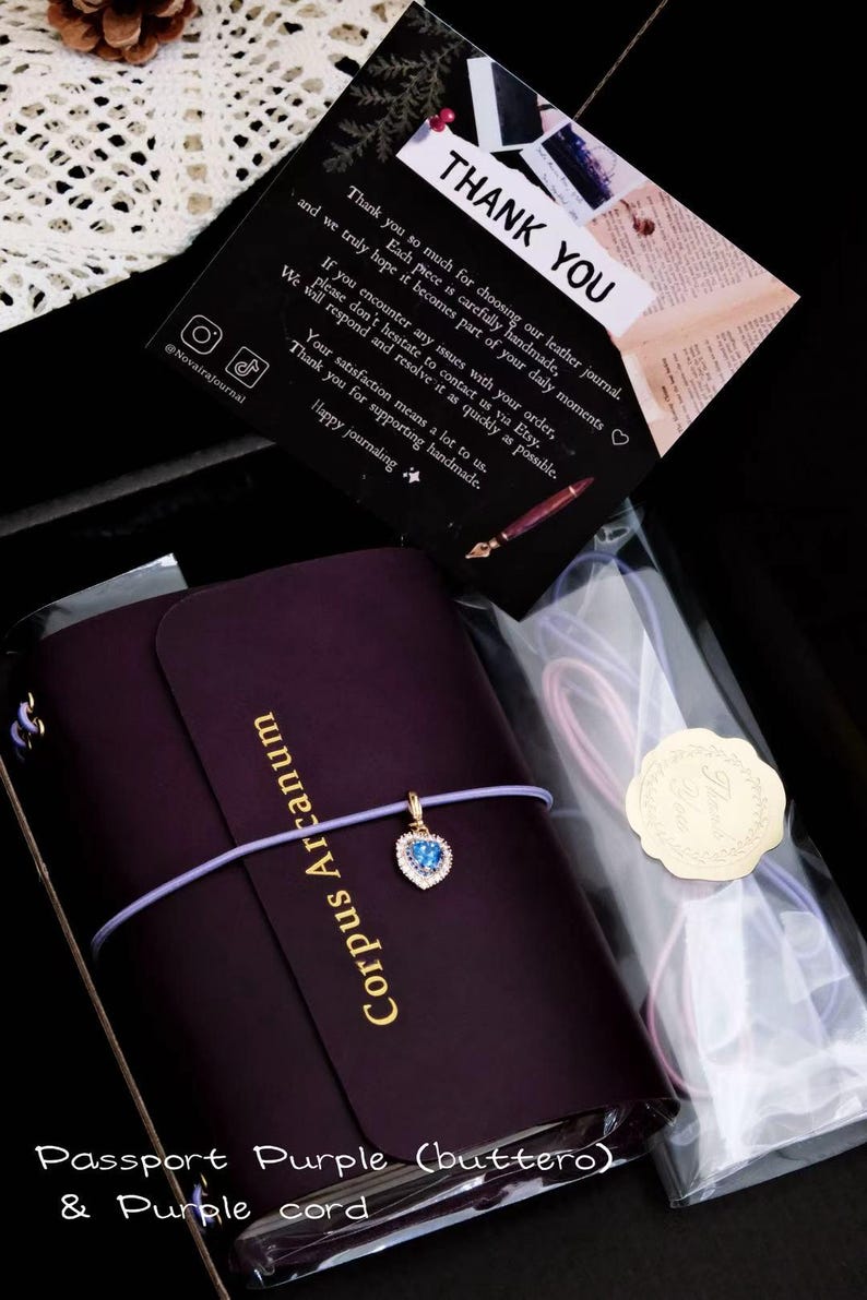 May include: A dark purple leather journal with the text Corpus Arcanum embossed in gold. A light purple elastic cord wraps around the journal, featuring a heart-shaped charm with a blue stone. A thank you card and extra cords are included.