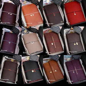 May include: A collection of twelve leather journals in various colours including burgundy, chestnut, foresta, red, purple, grey, brown, and black. Each journal features a coloured elastic band with a small metal charm and custom text.