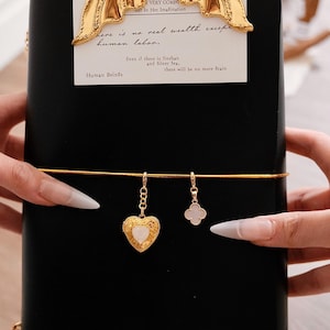 May include: A black notebook with a gold mermaid tail bookmark. The notebook has a gold elastic band and two gold charms: a heart-shaped locket and a four-leaf clover. Text on the page reads "Human Beings" and "Aria".