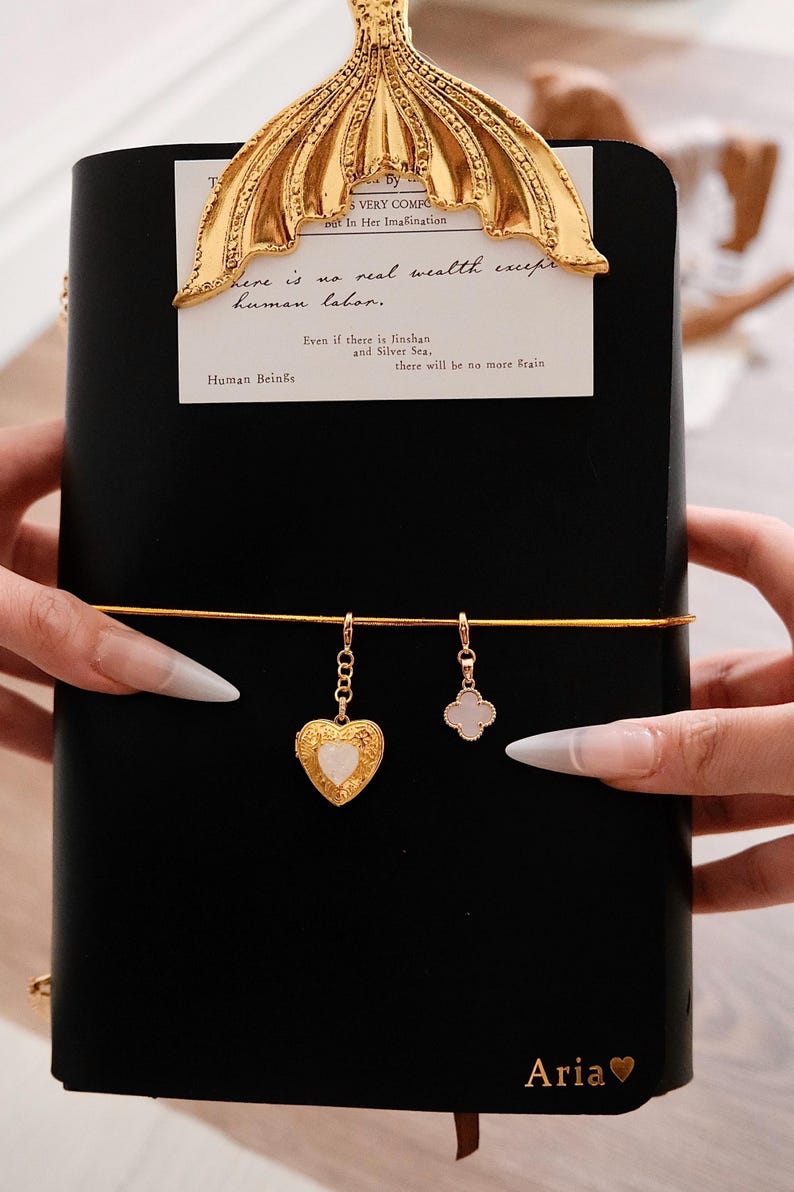May include: A black notebook with a gold mermaid tail bookmark. The notebook has a gold elastic band and two gold charms: a heart-shaped locket and a four-leaf clover. Text on the page reads "Human Beings" and "Aria".