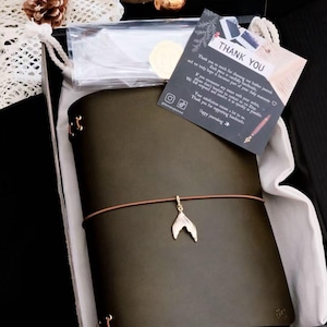 May include: An A5 olive green leather journal with a brown elastic cord featuring a gold-toned mermaid tail charm. The journal is presented in a black box with a thank you card that reads, "THANK YOU. Thank you so much for choosing our leather journal."