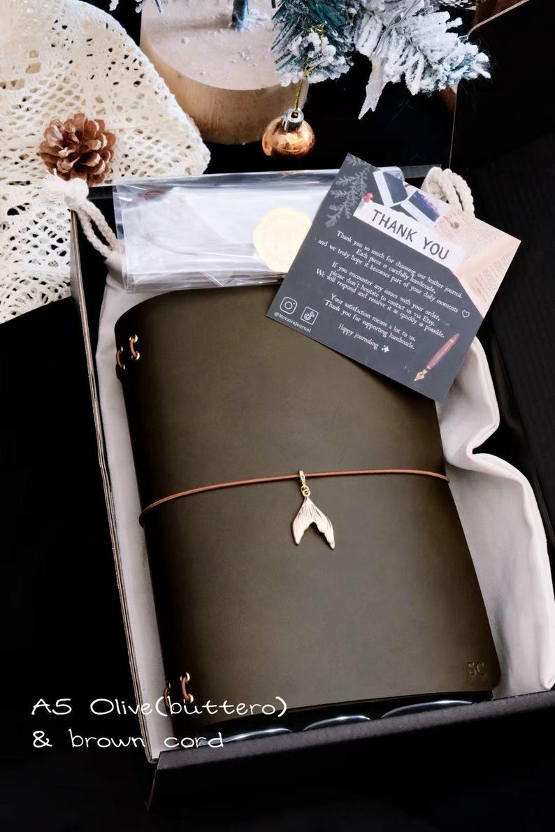 May include: An A5 olive green leather journal with a brown elastic cord featuring a gold-toned mermaid tail charm. The journal is presented in a black box with a thank you card that reads, "THANK YOU. Thank you so much for choosing our leather journal."