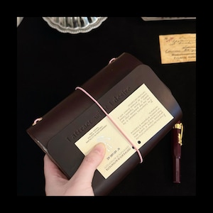 May include: A dark brown leather journal featuring the embossed text "I attract what I desire" on the front cover. The journal is secured with a thin pink elastic band and includes a cream-coloured certificate of authenticity card.