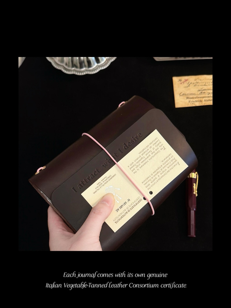 May include: A dark brown leather journal featuring the embossed text "I attract what I desire" on the front cover. The journal is secured with a thin pink elastic band and includes a cream-coloured certificate of authenticity card.