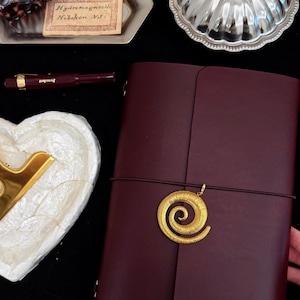 May include: A dark burgundy leather journal featuring a gold-toned spiral charm attached to an elastic closure band. The cover is embossed with the letters AP at the bottom. The journal is positioned alongside a burgundy fountain pen, a silver shell-shaped dish, and a small rectangular tray containing a tortoiseshell hair clip and a vintage label reading Hydromagnesite Hoboken NJ.