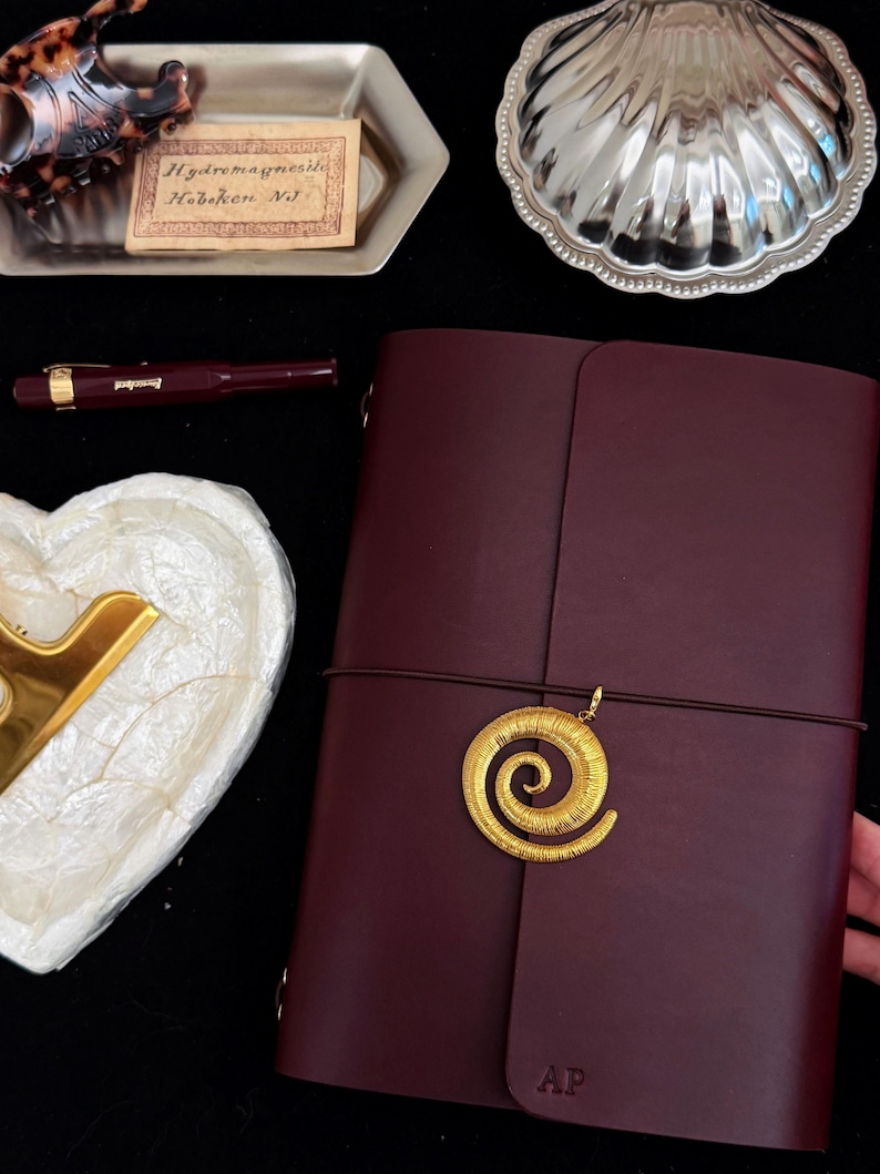 May include: A dark burgundy leather journal featuring a gold-toned spiral charm attached to an elastic closure band. The cover is embossed with the letters AP at the bottom. The journal is positioned alongside a burgundy fountain pen, a silver shell-shaped dish, and a small rectangular tray containing a tortoiseshell hair clip and a vintage label reading Hydromagnesite Hoboken NJ.