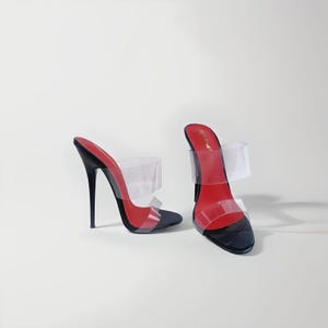 Könnte beinhalten: Ein Paar schwarze High-Heel-Mules mit transparenten Riemen. Die Schuhe haben eine rote Innensohle und eine spitze Zehenpartie. Die Absätze sind etwa 10 cm hoch. Die Schuhe stehen vor weißem Hintergrund.
