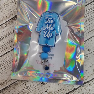 May include: A blue badge reel with a lab coat design and the text "Tie Me Up". The badge reel has blue beads and a clear plastic clip. It is packaged in a holographic zip bag.