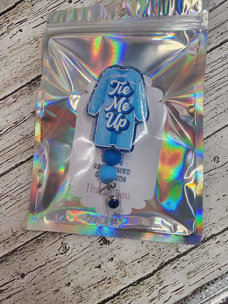 May include: A blue badge reel with a lab coat design and the text "Tie Me Up". The badge reel has blue beads and a clear plastic clip. It is packaged in a holographic zip bag.