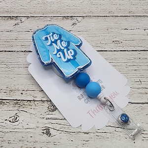 May include: A blue badge reel with a scrub top design and the words "Tie Me Up". The reel has two blue beads and a clear plastic clip with a blue glitter accent. The badge is on a white card.