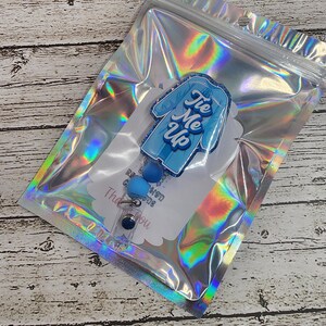 May include: A blue and white badge reel with the text "Tie Me Up" in a script font. The badge reel is in a clear, holographic bag. The badge reel has a blue, round button and a clear plastic clip.