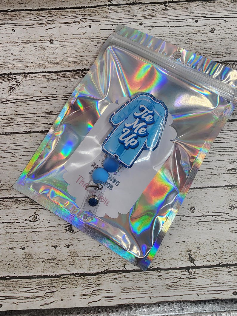 May include: A blue and white badge reel with the text "Tie Me Up" in a script font. The badge reel is in a clear, holographic bag. The badge reel has a blue, round button and a clear plastic clip.
