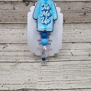 May include: A light blue badge reel with a medical coat design and the words "Tie Me Up" in cursive. The badge reel has blue beads and a clear plastic clip. The background is a light wood grain.