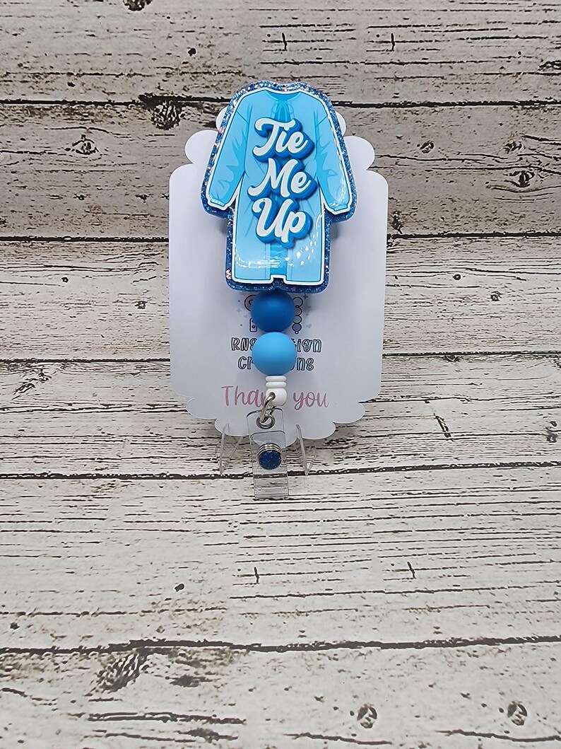 May include: A light blue badge reel with a medical coat design and the words "Tie Me Up" in cursive. The badge reel has blue beads and a clear plastic clip. The background is a light wood grain.