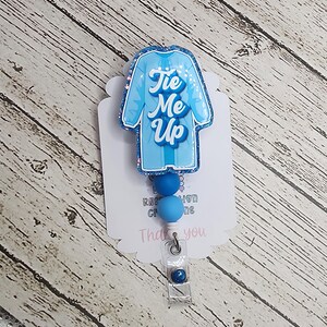 May include: A light blue badge reel with a glittery outline, shaped like a jersey, featuring the text "Tie Me Up" in a script font. The badge reel has blue beads and a clear plastic clip.