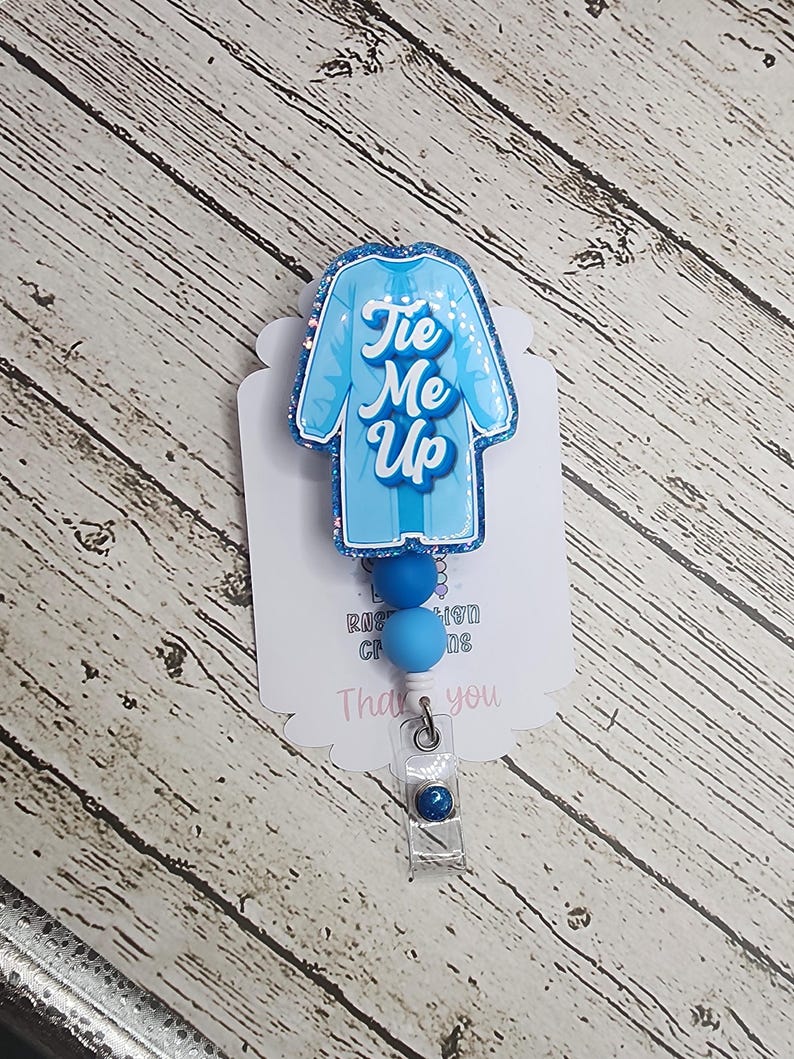 May include: A light blue badge reel with a glittery outline, shaped like a jersey, featuring the text "Tie Me Up" in a script font. The badge reel has blue beads and a clear plastic clip.