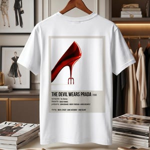 May include: White t-shirt featuring a graphic from "The Devil Wears Prada" movie. The design includes a red high heel with a trident heel and the movie title, year, and credits. The shirt is hanging on a wooden hanger.