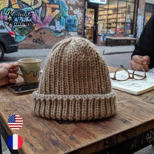 May include: A tan knitted beanie hat with a ribbed brim sits on a wooden table. The hat is made of thick yarn and has a textured appearance. The words "Knitting Pattern" are in a circle in the upper left corner.