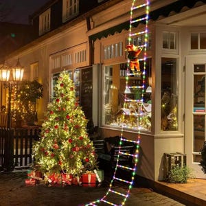 Puede incluir: Una escena navideña con un árbol decorado con adornos rojos y regalos. Una escalera de luces multicolores se extiende desde el techo, con una figura de Papá Noel que la escala. La escena está ambientada frente a un edificio con ventanas iluminadas.