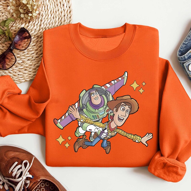 Może przedstawiać: Pomarańczowa bluza z okrągłym dekoltem z grafiką Buzza Astrala i Chudego z Toy Story. Buzz ma na sobie skafander kosmiczny ze skrzydłami, a Chudy jest w stroju kowbojskim. Projekt zawiera ż&oacute;łte gwiazdki.