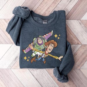 Peut inclure: Un sweat-shirt gris foncé avec un motif de Buzz l'Éclair et Woody de Toy Story. Buzz porte sa combinaison spatiale avec des ailes, et Woody est en tenue de cow-boy. Le motif comprend des étoiles jaunes.