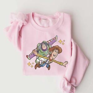Może przedstawiać: Jasnor&oacute;żowa bluza z kresk&oacute;wką Buzza Astrala i Chudego z Toy Story. Buzz ma na sobie skafander kosmiczny, a Chudy str&oacute;j kowbojski. Projekt zawiera ż&oacute;łte gwiazdy.