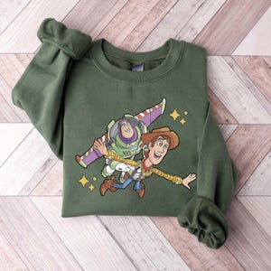Może przedstawiać: Zielona bluza z grafiką Buzza Astrala i Chudego z Toy Story. Postacie z kresk&oacute;wek są przedstawione w powietrzu z gwiezdnym tłem. Bluza ma długie rękawy i okrągły dekolt.
