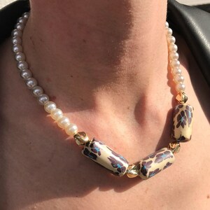 Peut inclure: Un collier de perles avec trois perles cylindriques crème ornées d'un motif floral bleu et marron. Le collier a des accents dorés et est porté sur une veste en cuir noire.