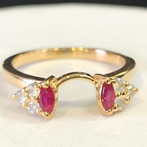 May include: A gold-colored ring with a unique design. The ring features two marquise-cut, deep red gemstones and several smaller, clear gemstones. The ring's design is open, allowing it to wrap around another ring.
