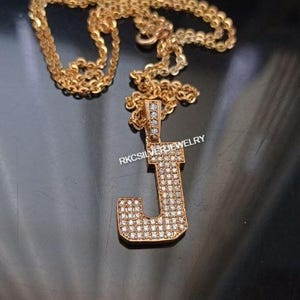 May include: Gold-colored necklace with a pendant in the shape of the letter "J". The pendant is encrusted with small, clear gemstones. The chain is a delicate link style. The text "RKCSILVERJEWELRY" is visible on the pendant.